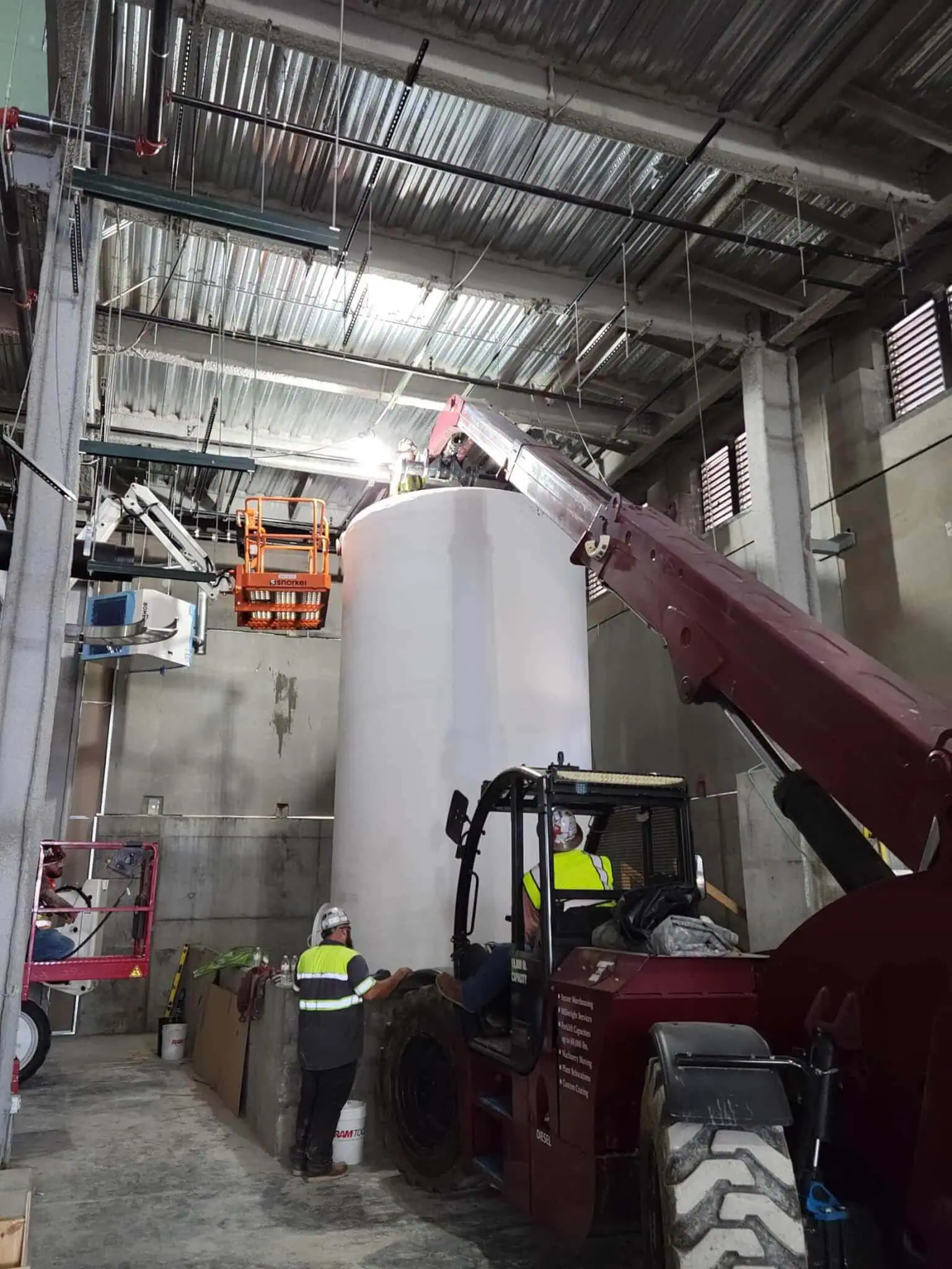MEI Industrial Solutions crew operating a Versa-Lift forklift with boom to lift a large fiberglass waste tank indoors