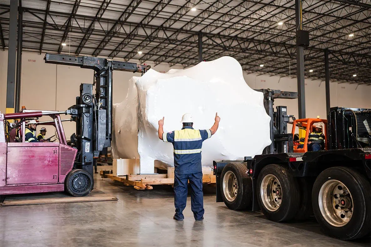 MEI Industrial Solutions riggers and signal person using dual heavy-duty forklifts to load a large shrink-wrapped industrial machine onto a flatbed trailer indoors.